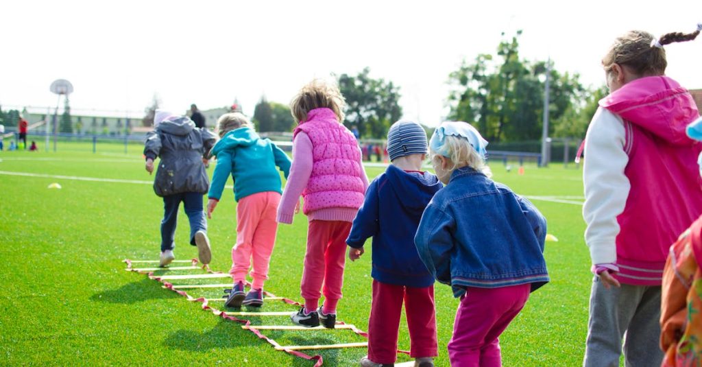 Children enjoy an outdoor activity on a grassy field, stepping over a ladder.